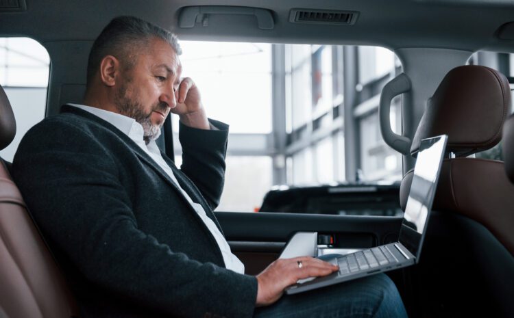 Side view. Working on a back of car using silver colored laptop. Senior businessman.